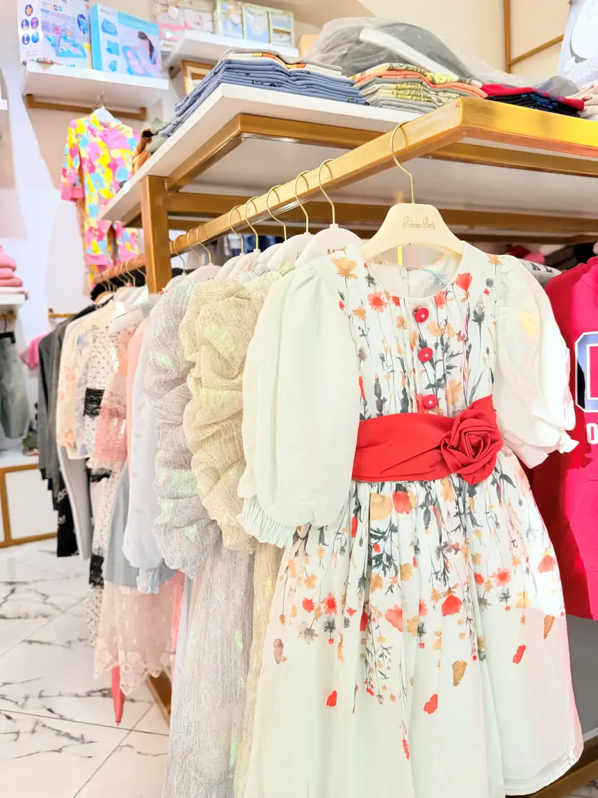 A white flowered party dress with a red rose sash on a gold rail at Sultan Kids